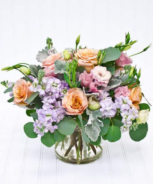 A classic pastel color pallet perfect for Spring! Peach roses are beautifully combined with pink lisianthus and lavender stock all organically designed with eucalyptus and dusty miller foliage. Overall arrangement is approximately 12"H x 14"W and perfect for a dining room table or large kitchen island.
