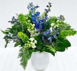 An impressive display of blue delphinium, white snapdragon, and green bells of Ireland is designed in an elegant white glass vase and accented with fragrant cedar.